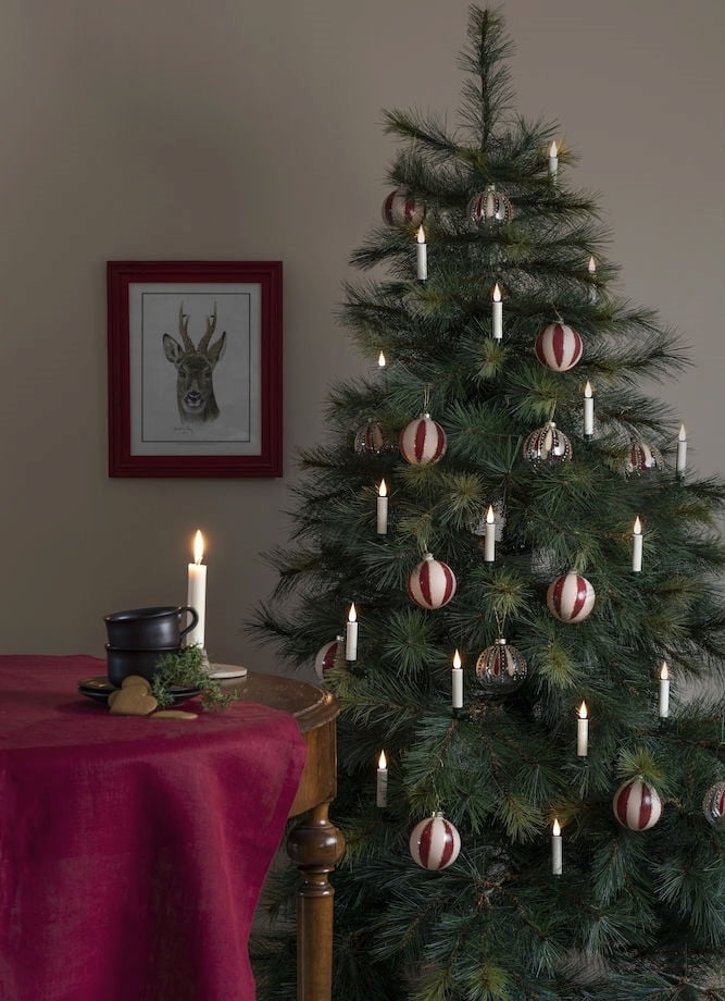 A cozy Christmas scene with a decorated pine tree, striped ornaments, white candles, and a red tablecloth beside a framed deer sketch.