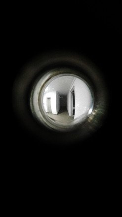 Peephole view into a brightly lit hallway with white walls and doors, seen through a fisheye lens effect.