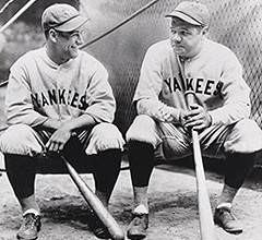 Two New York Yankees baseball players in vintage uniforms crouching with bats, smiling on a field.