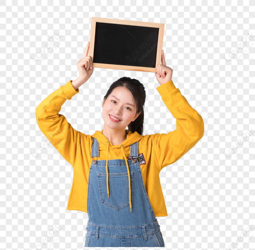 Young woman in yellow hoodie and denim overalls holding a blank chalkboard above her head, smiling at the camera.