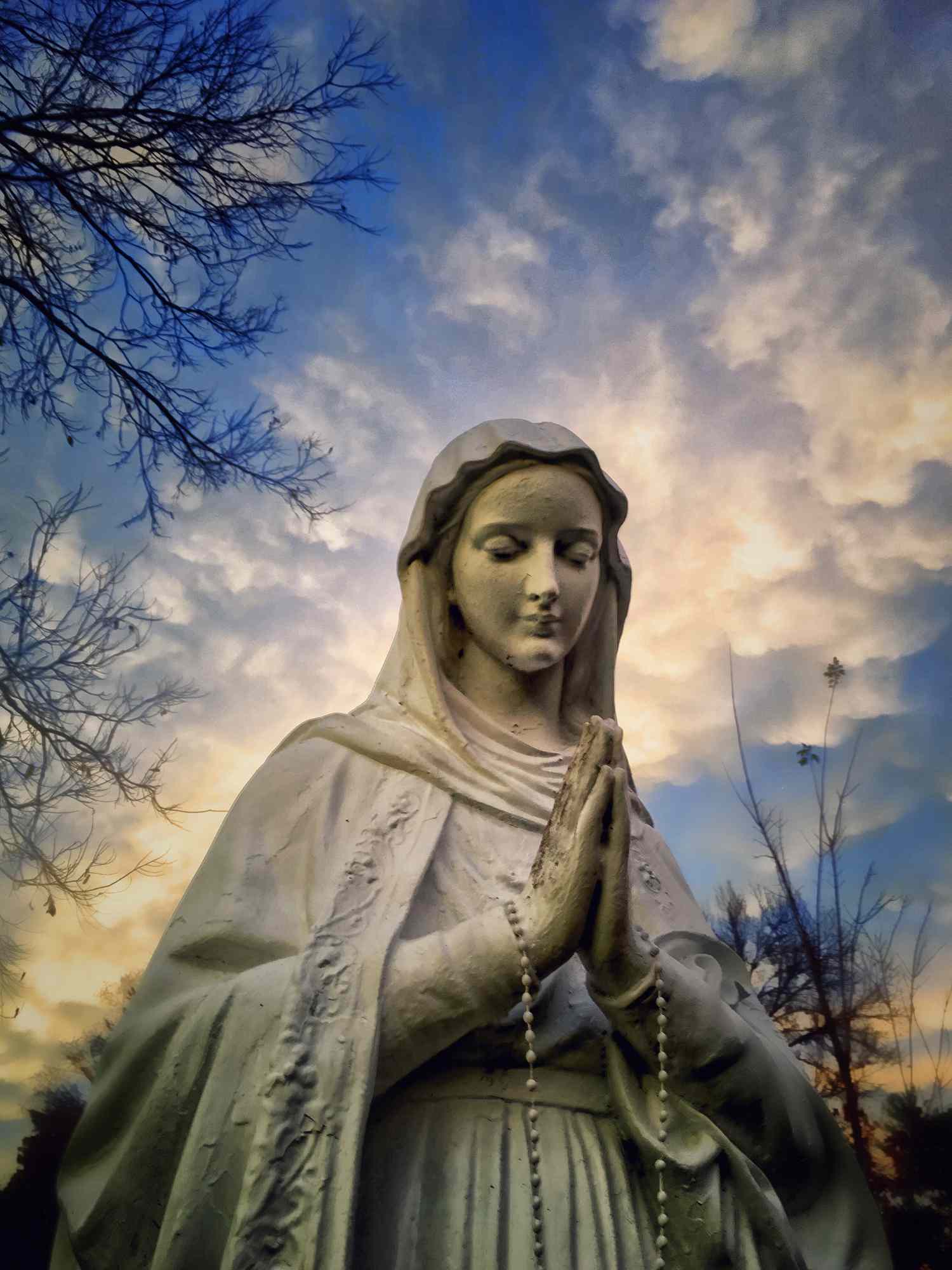 Stone statue of the Virgin Mary in prayer, hands clasped with rosary, against a dramatic cloudy sky and bare tree branches.