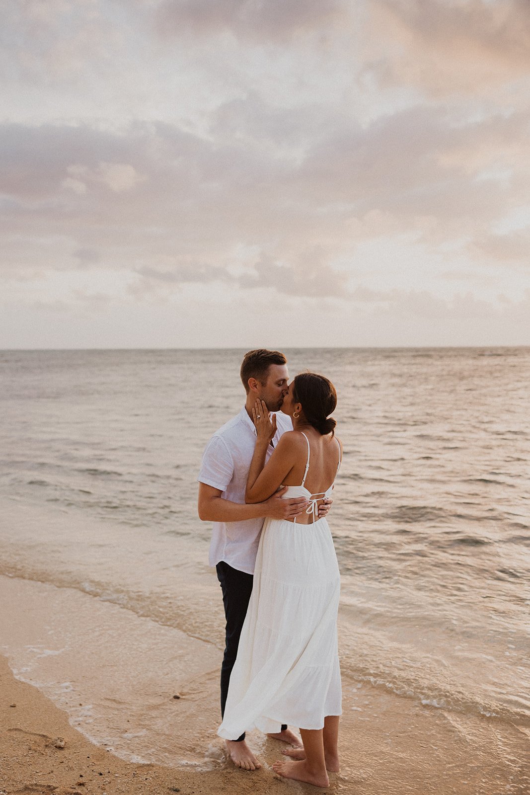 A couple shares a tender kiss on a sandy beach at sunset, wearing white outfits with gentle waves lapping at their feet.