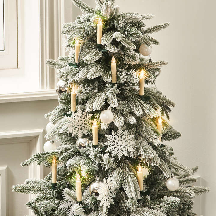 Flocked Christmas tree decorated with white snowflakes, pearl ornaments, and warm LED candle lights near a window.
