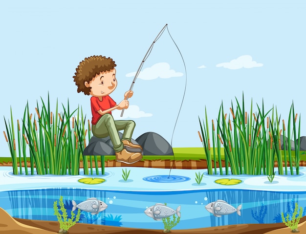 A smiling boy in a red shirt sits on a rock by a pond, fishing with a rod while fish swim below the surface among reeds and lily pads.