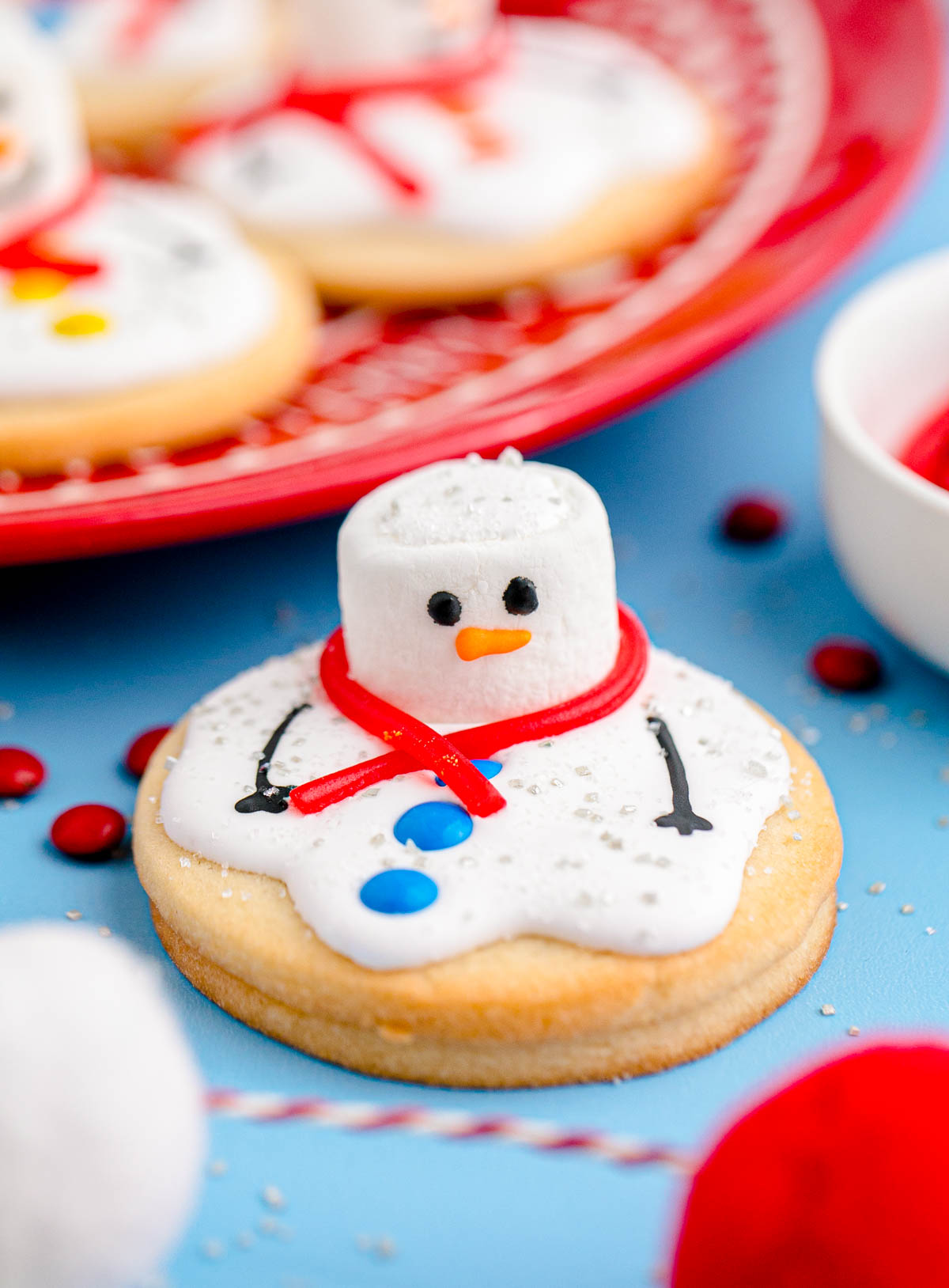 Snowman cookie with marshmallow head, red licorice scarf, blue candy buttons, and black icing details on a blue surface.