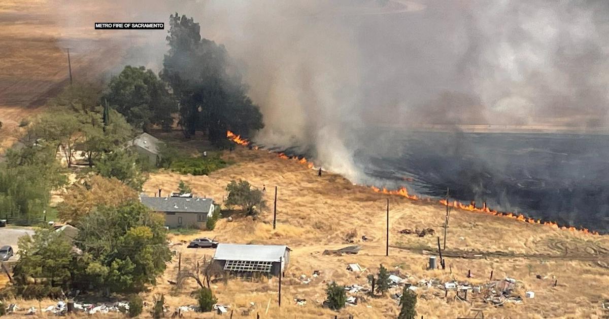 Aerial view of the Metro Fire of Sacramento burning through dry grassland near homes and structures, with thick smoke rising.