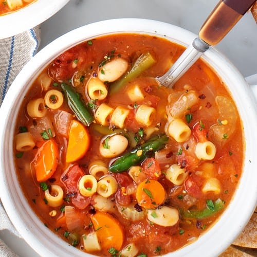 Hearty vegetable minestrone soup with pasta, carrots, green beans, tomatoes, and herbs in a white bowl with spoon.