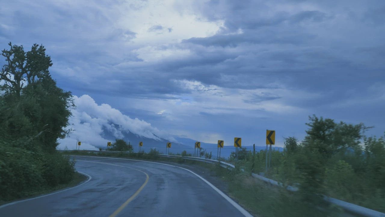 Winding mountain road with yellow curve warning signs under dramatic cloudy sky and distant peaks.