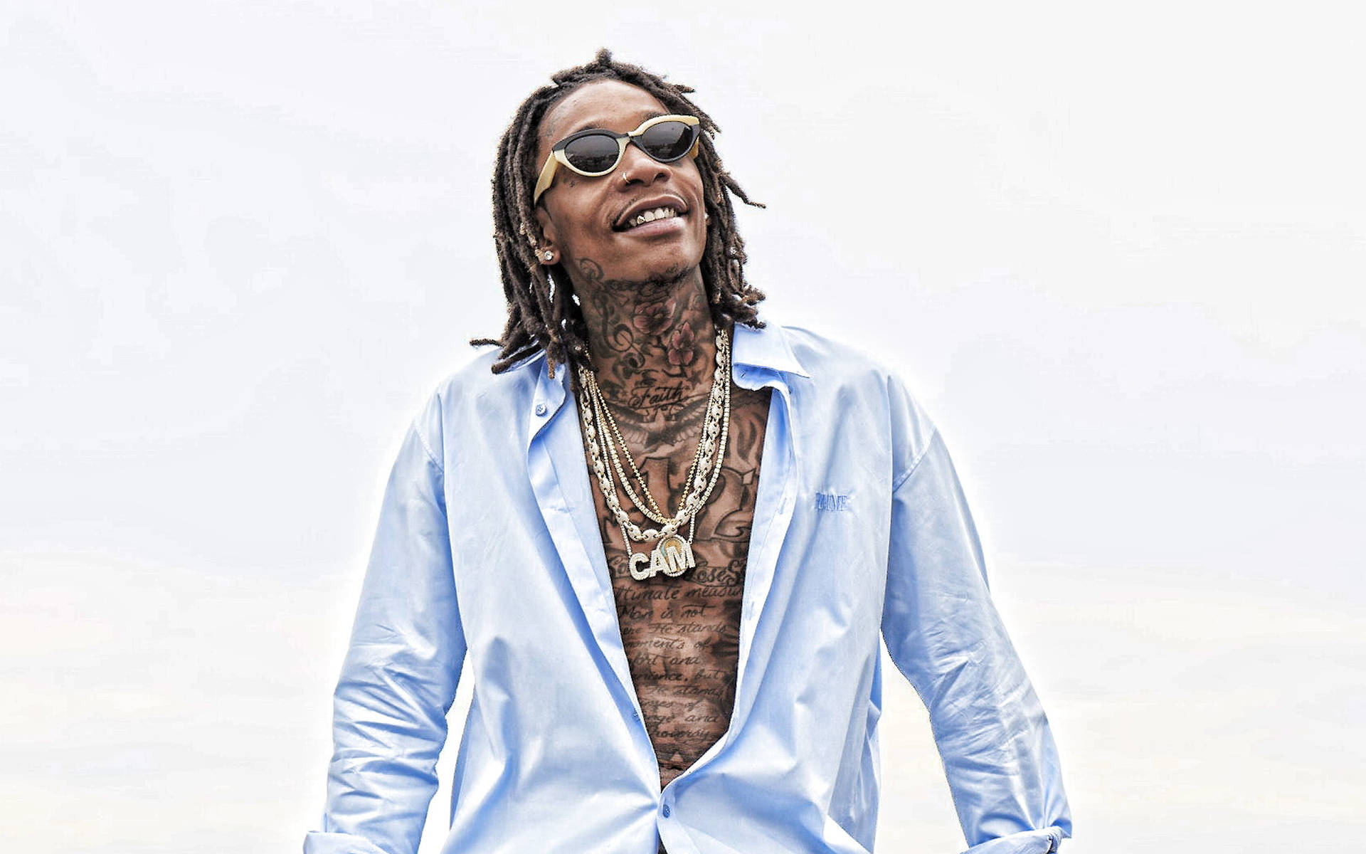 Rapper with dreadlocks, sunglasses, tattoos, and layered gold chains wearing an open light blue shirt against a bright background.