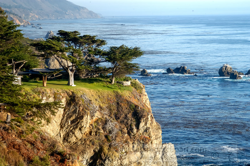 Coastal cliff with cypress trees, modern house, and Pacific Ocean view near Big Sur, California.