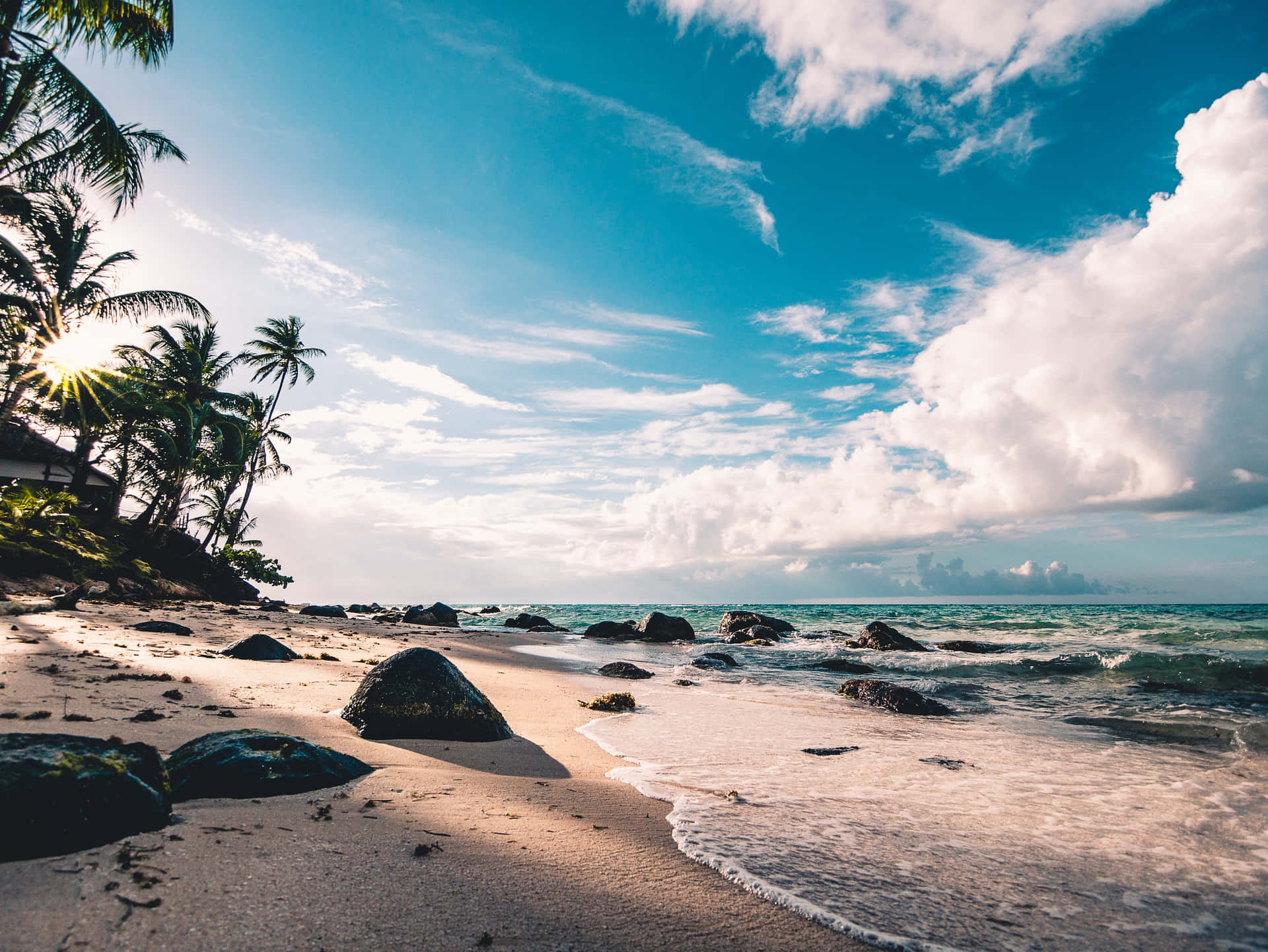 Serene tropical beach with palm trees, golden sand, scattered rocks, gentle waves, and sunburst through clouds at dawn.