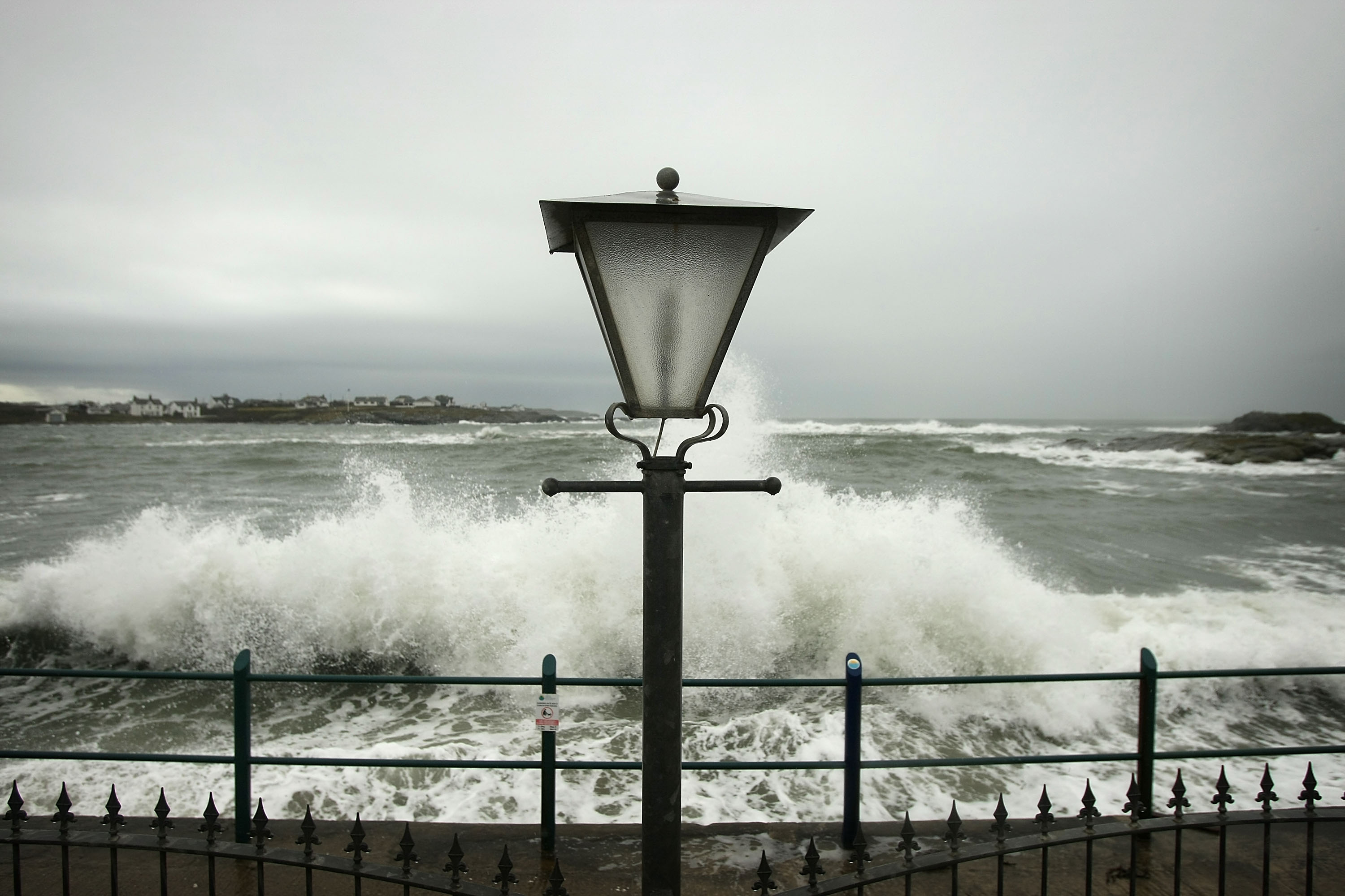 A vintage streetlamp stands before crashing waves on a stormy coastal promenade with distant houses under grey skies.