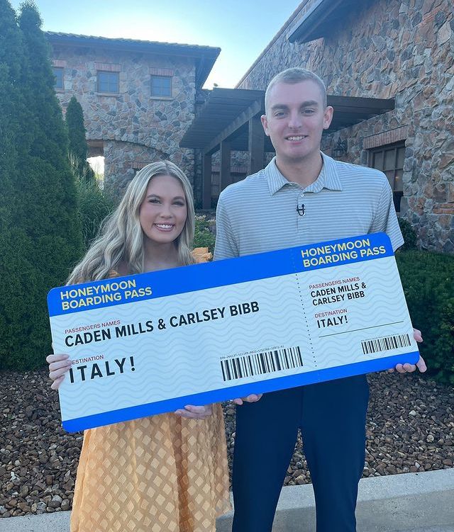 A smiling couple holds a giant "Honeymoon Boarding Pass" to Italy, standing in front of a stone building.