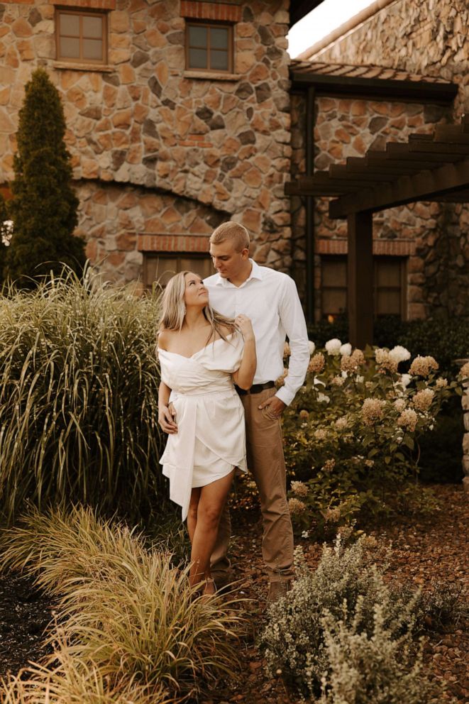 A couple poses affectionately in a garden beside a stone building, woman in white dress, man in white shirt and khakis.