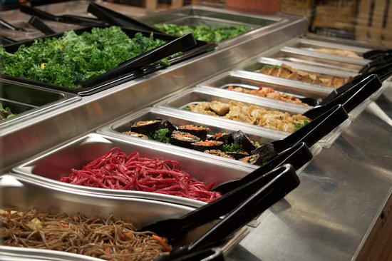 Stainless steel buffet trays filled with colorful fresh foods including kale, pickled ginger, sushi, and stir-fried noodles.