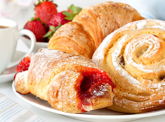 Freshly baked pastries including croissants, a strawberry-filled pastry, and a cinnamon swirl, dusted with powdered sugar, beside strawberries and coffee.