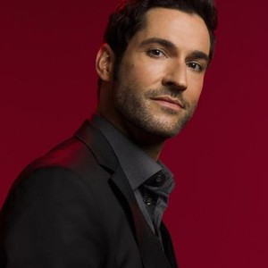 Tom Ellis in a dark suit against a deep red background, looking confidently at the camera.