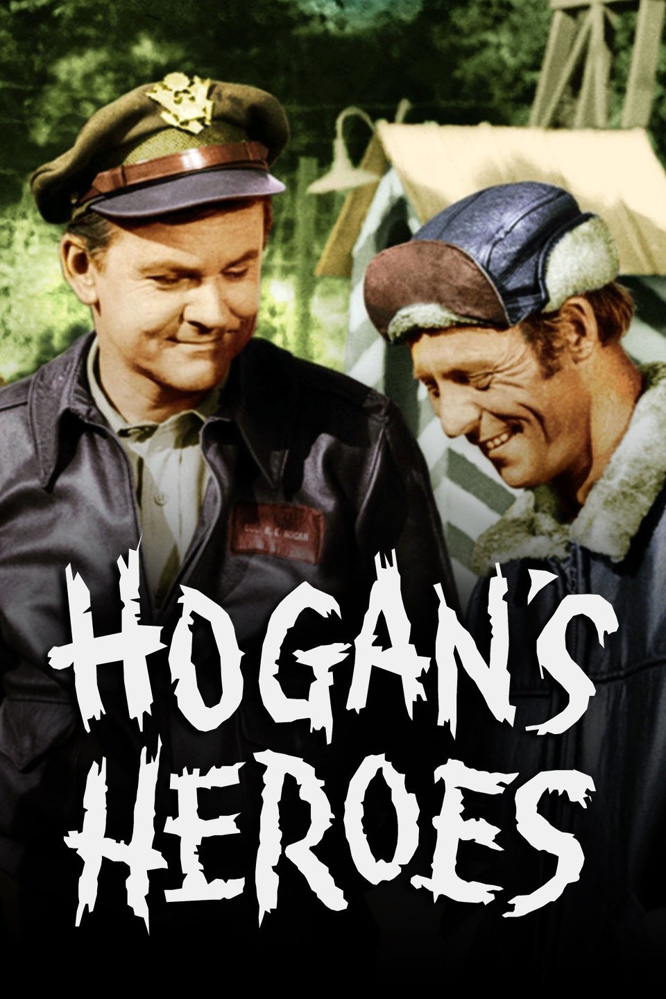 Two smiling WWII-era soldiers in military uniforms, one wearing a cap with insignia and the other a fur-lined aviator hat, outdoors near tents.