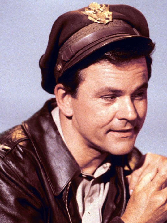 Actor in WWII-era pilot uniform with leather jacket and cap, smiling subtly.