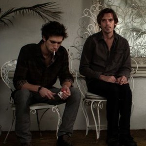 Two men seated on ornate white chairs in a softly lit interior, one holding a cigarette and a small object, the other gazing forward pensively.