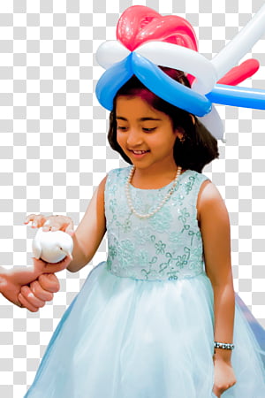 A smiling girl in a light blue dress wearing a colorful balloon hat, holding a small white plush toy handed by an adult.