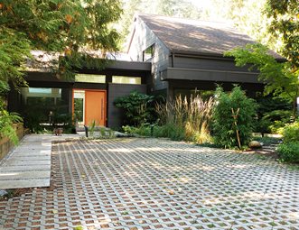 Modern single-story home with dark exterior, orange door, and permeable paver driveway surrounded by lush greenery.