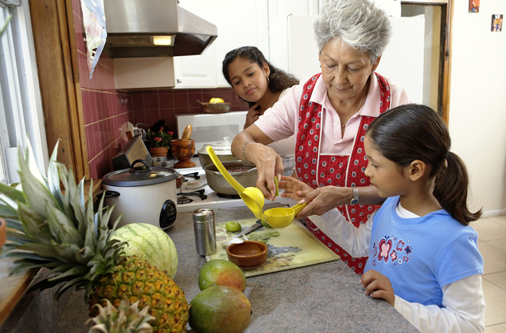Elderly woman and two girls preparing fruit in a home kitchen, using a citrus juicer.