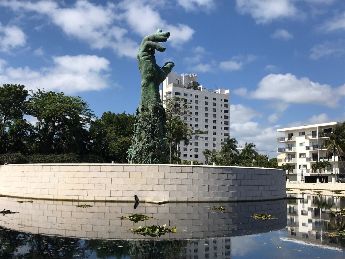 A large bronze sculpture of a hand emerging from water, surrounded by a circular stone wall and reflecting pool, with modern buildings in background.