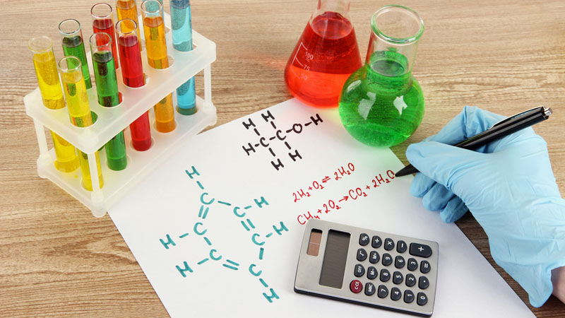 Scientist in blue gloves writes chemical formulas on paper beside test tubes, beakers, and a calculator on a wooden lab table.