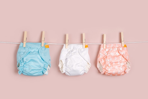 Three colorful reusable cloth diapers-blue, white, and peach with patterns-hanging on a clothesline against a soft pink background.