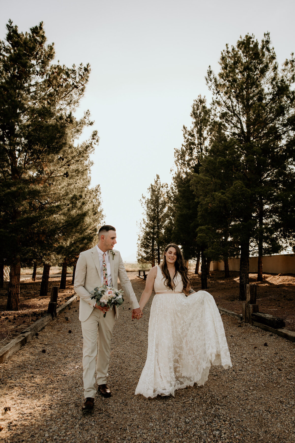 A bride and groom walk hand-in-hand down a gravel path lined with tall pine trees, smiling warmly in golden-hour light.