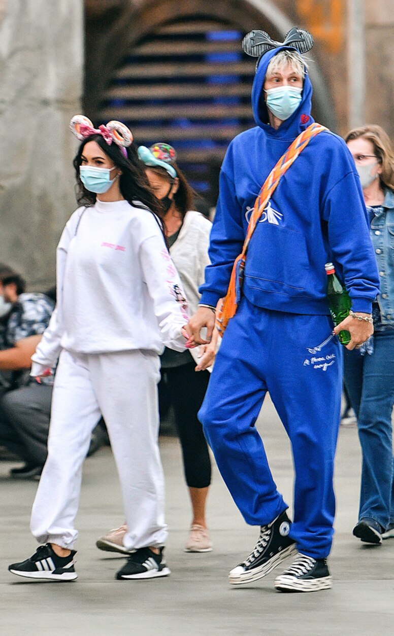 A couple wearing Disney-themed outfits and face masks walk hand-in-hand, the man in a blue hoodie with Mickey ears and the woman in white with donut ears.