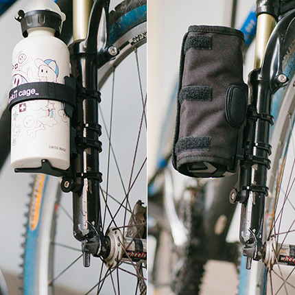Two bicycle bottle cages mounted on a fork: one holding a white illustrated water bottle, the other a black fabric sleeve-style cage.