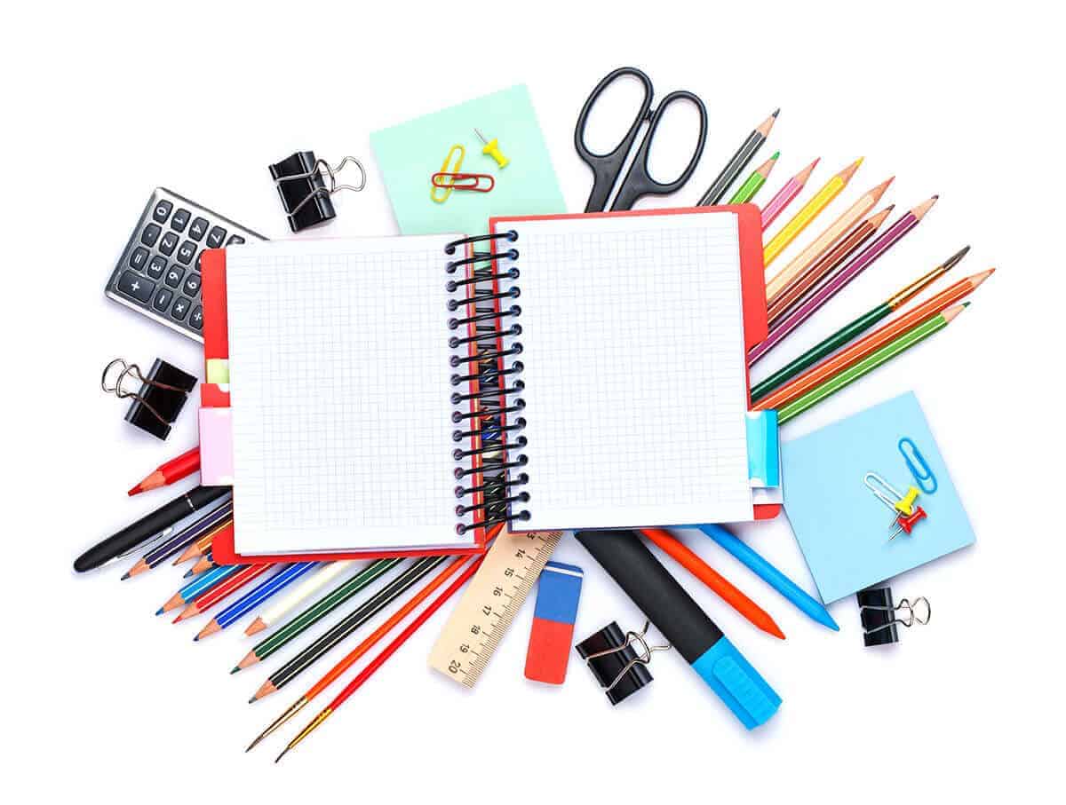 A colorful assortment of school and office supplies including a notebook, pencils, scissors, calculator, eraser, ruler, and paper clips on white background.