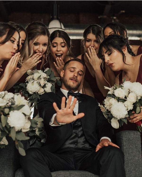 Groom in black tuxedo playfully raises hand as bridesmaids in maroon dresses react with surprise and laughter, holding white bouquets.