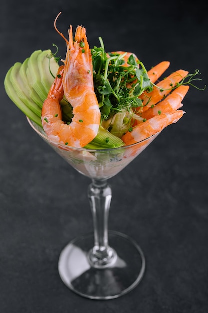 Shrimp cocktail in a martini glass with avocado, celery, and microgreens on dark background.