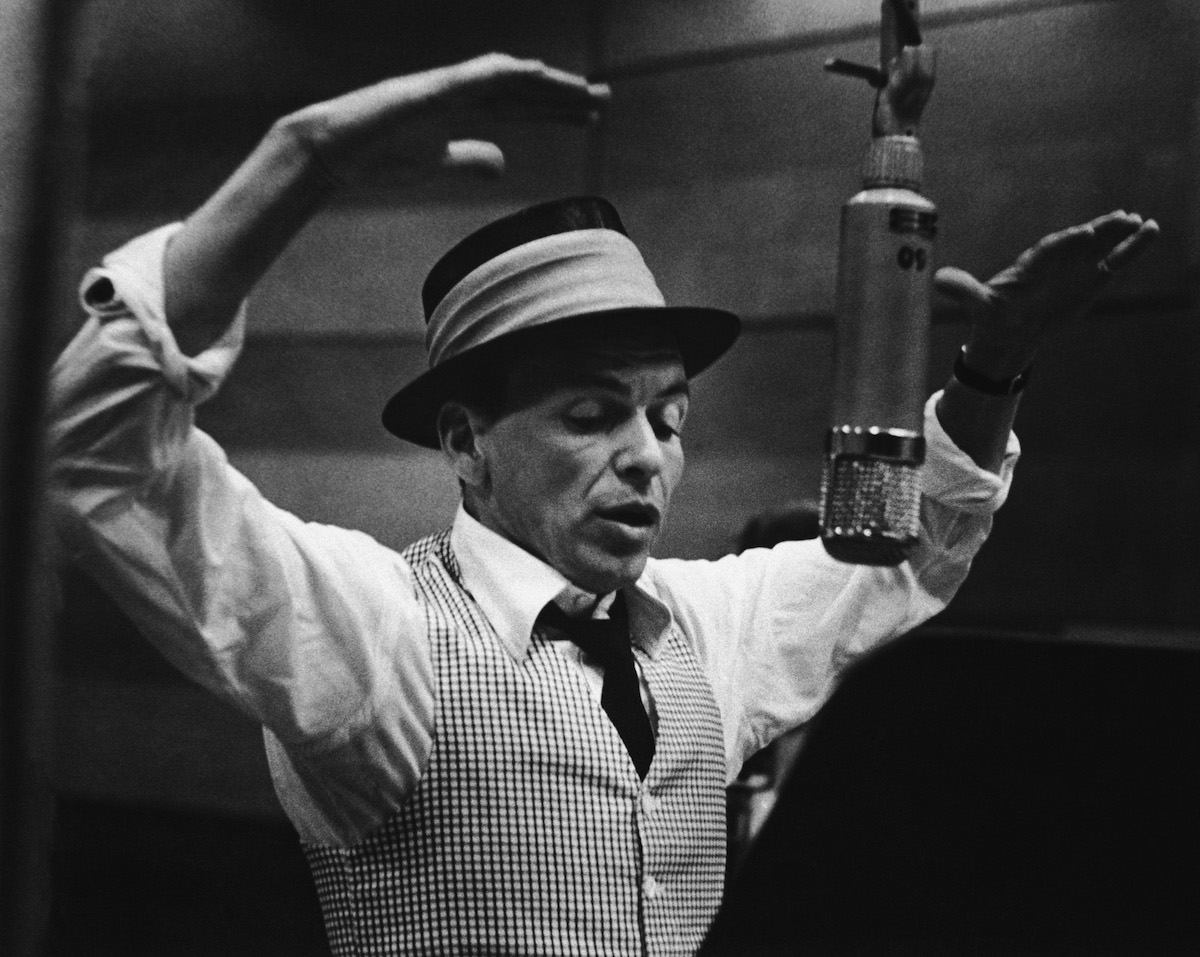 Frank Sinatra singing into a vintage microphone in a recording studio, wearing a fedora and checkered vest.