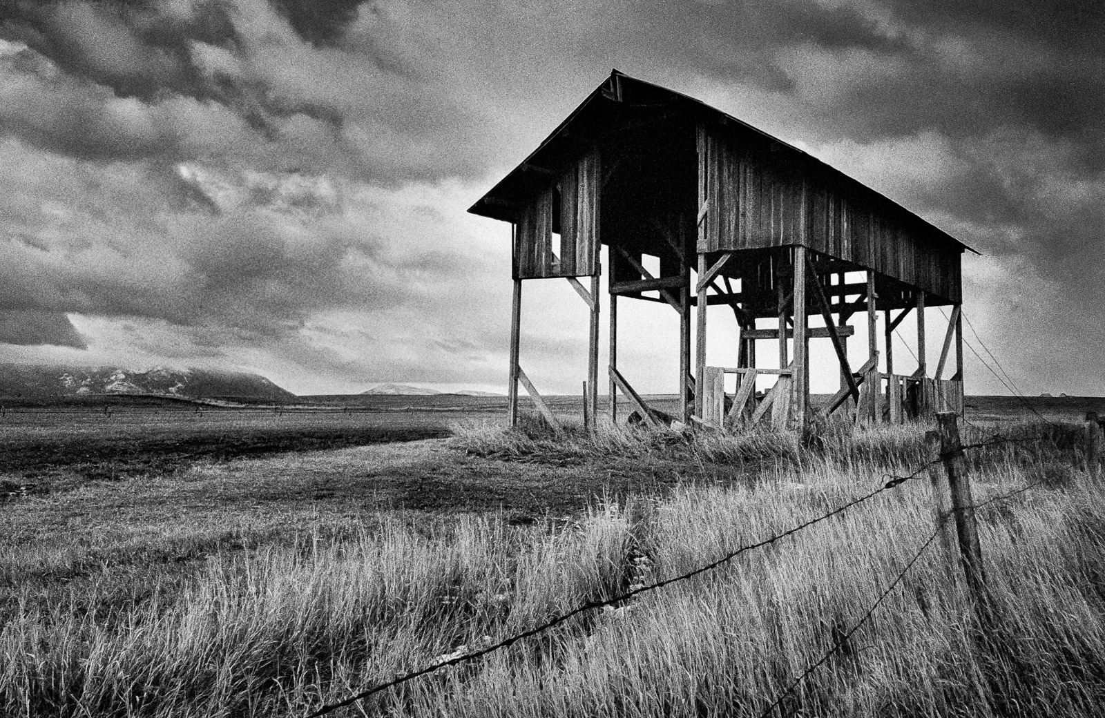 Weathered wooden hay barn on stilts in an open field under dramatic clouds, black-and-white landscape photo.