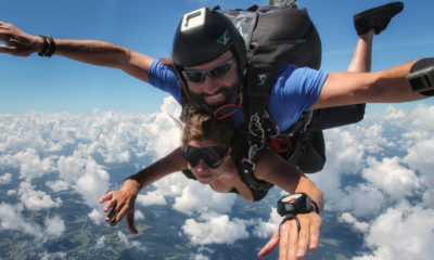 Tandem skydivers in freefall above clouds, instructor guiding student with arms outstretched.