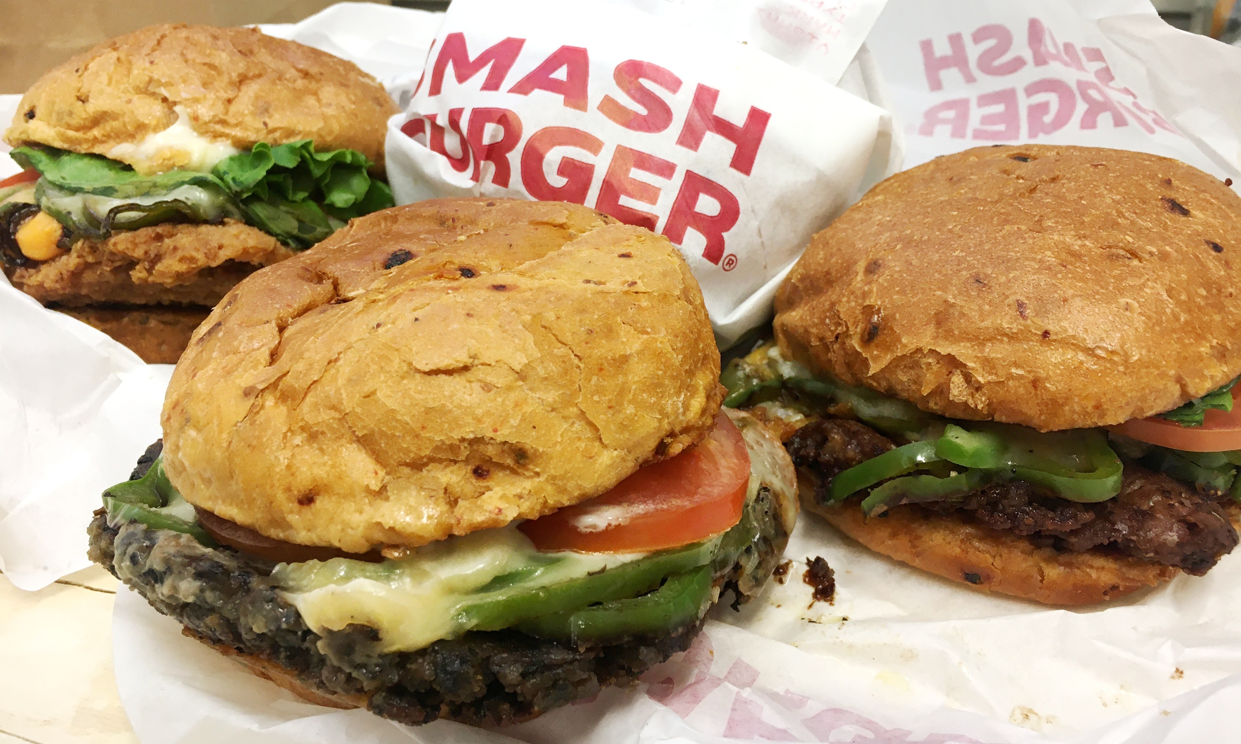 Three appetizing Mash Burger sandwiches with crispy buns, melted cheese, fresh veggies, and savory patties on branded paper wrapping.
