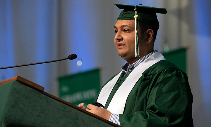 A graduate in green cap and gown delivers a speech at a podium during a commencement ceremony.