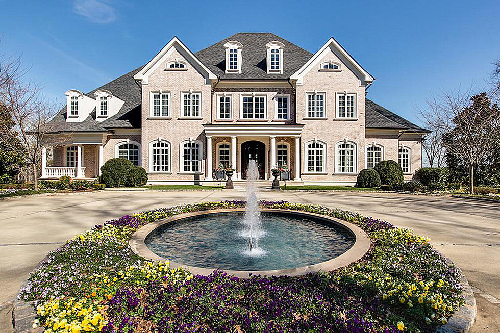 Grand two-story brick mansion with arched windows, columned entrance, and circular fountain surrounded by colorful flowerbeds.