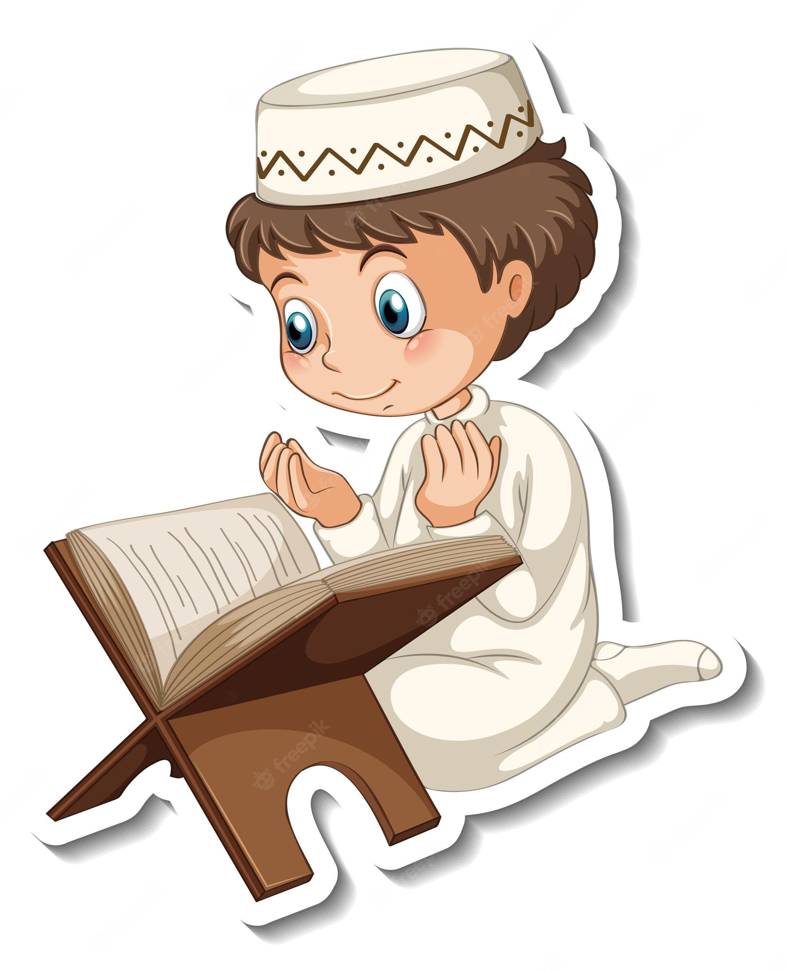 A smiling Muslim boy in a white thobe and kufi cap kneels while reading the Quran on a wooden stand.