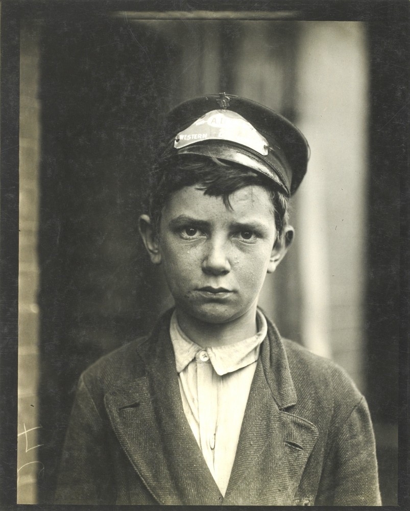 Young boy in early 20th-century Western Union cap and worn suit, solemn expression, vintage black-and-white portrait.