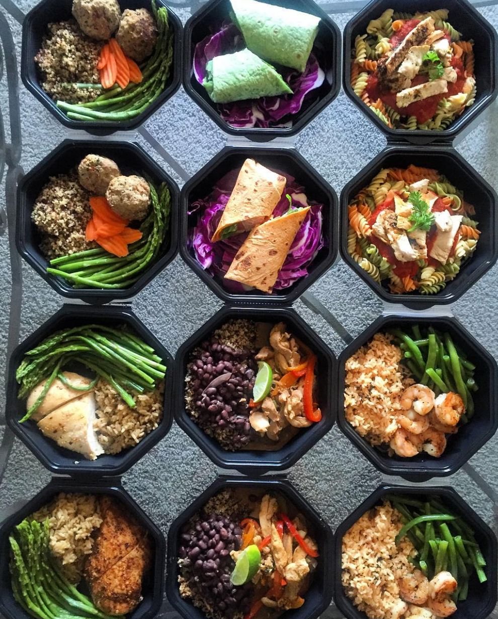 Twelve prepped healthy meals in black octagonal containers, featuring quinoa, chicken, shrimp, beans, asparagus, wraps, and pasta salads.