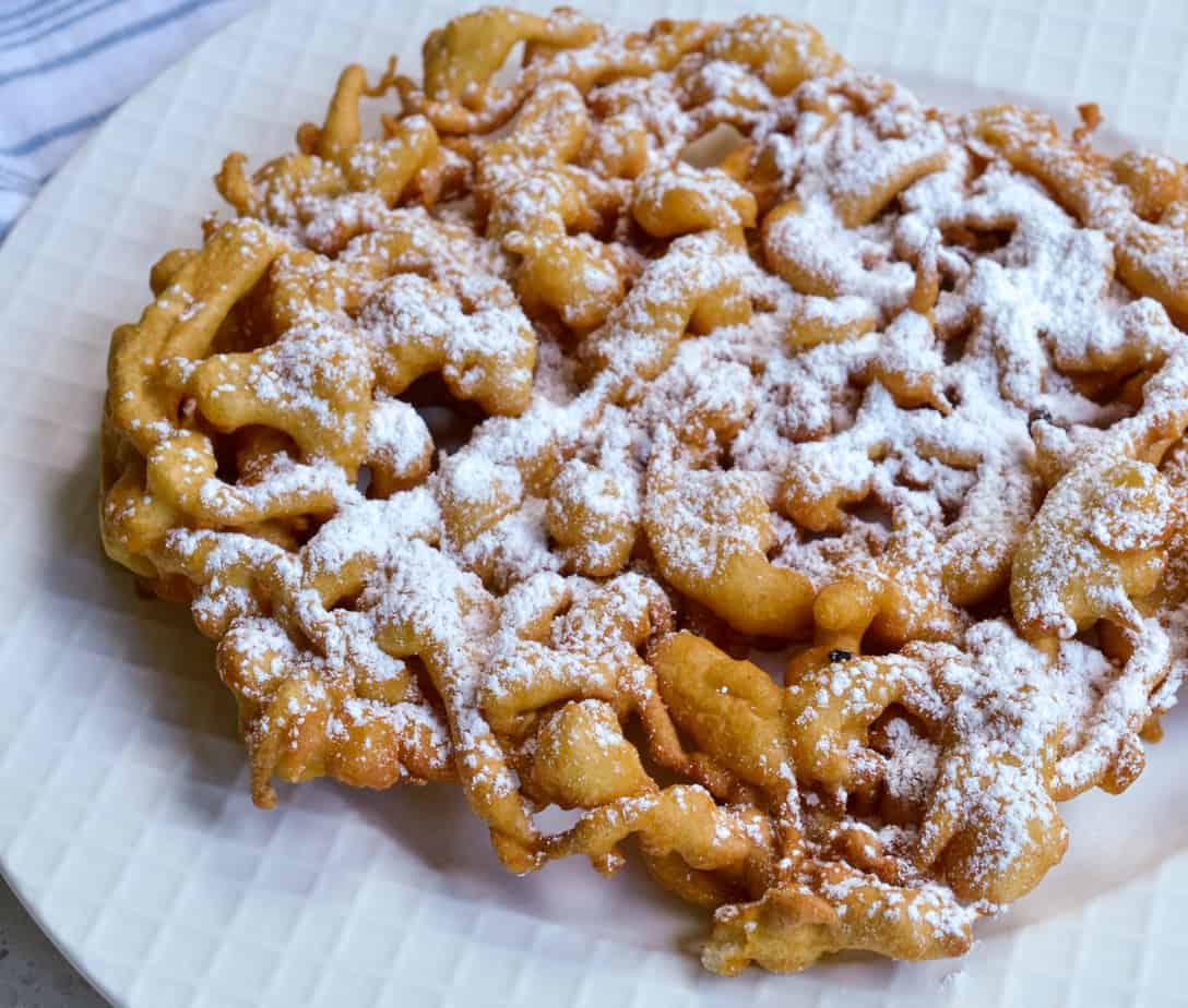 Golden, lacy funnel cake dusted with powdered sugar on a white plate.