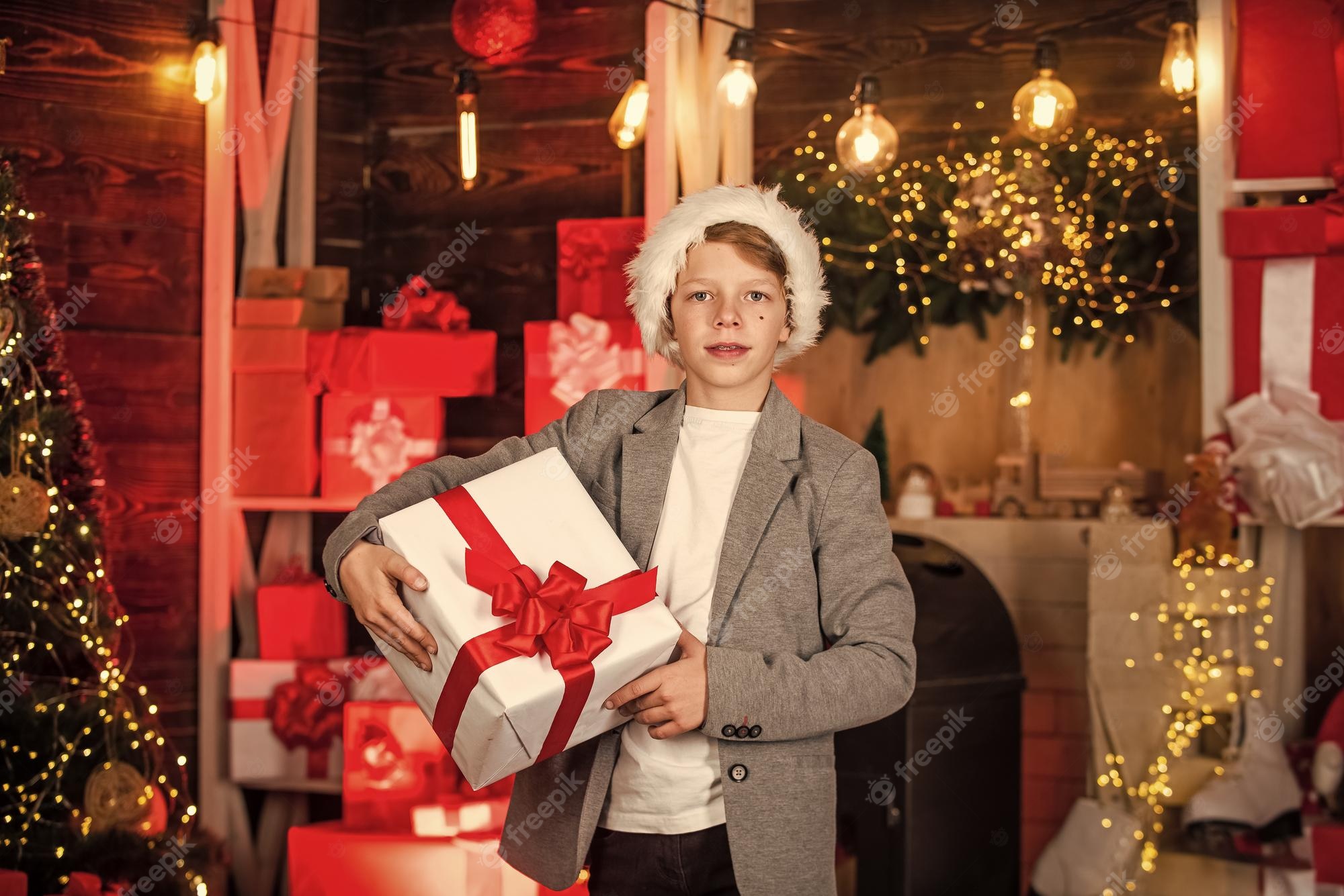 Boy in Santa hat holding a white gift box with red ribbon in a warmly lit Christmas-decorated room.