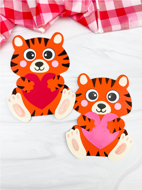 Two cute paper tiger crafts holding red and pink hearts on a white surface with a plaid fabric background.