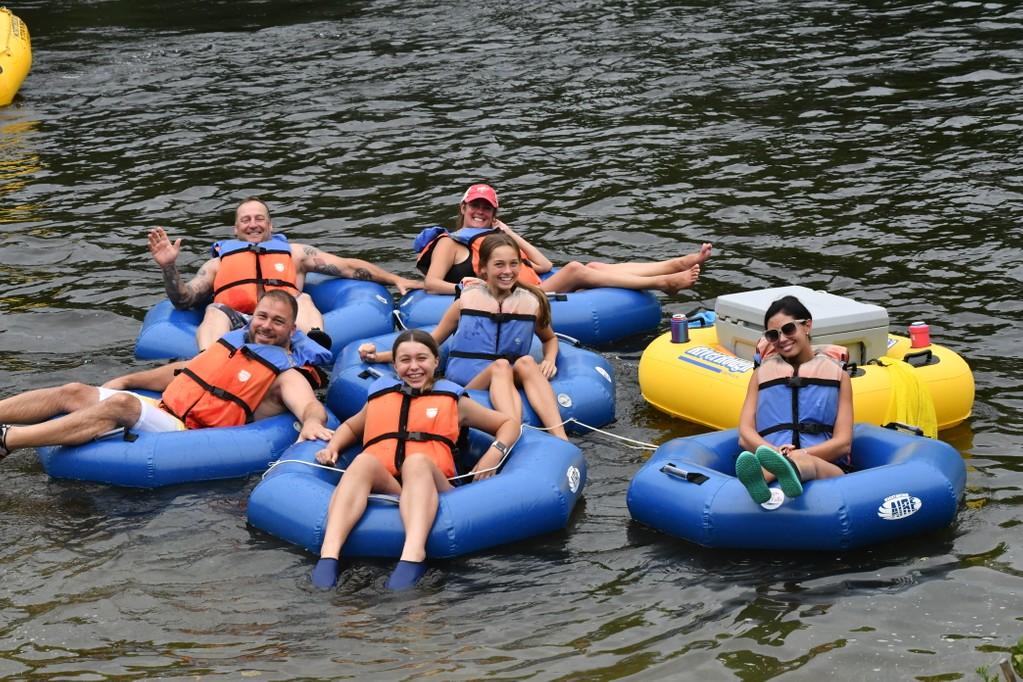 Group of six smiling adults tubing on a river in blue and yellow inflatable rafts with life jackets.