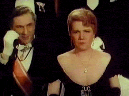 A formally dressed man in a tuxedo with sash and white gloves stands beside a woman in an elegant black gown with plunging neckline and pendant necklace.